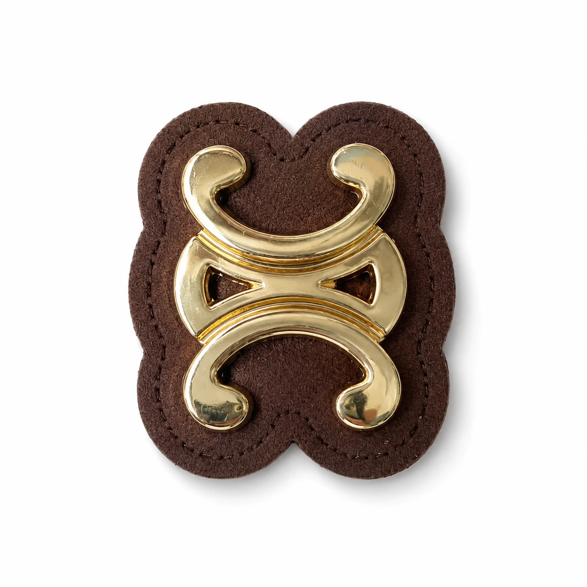 Front view of a gold-tone symmetrical scroll metal ornament on a brown stitched backing patch